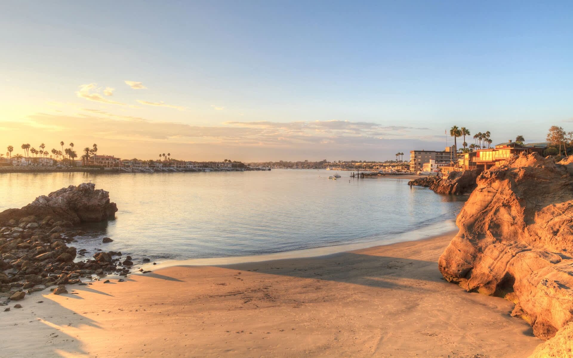 A calm channel at sunset, palms and houses along the far shore, sandstone rocks in the foreground